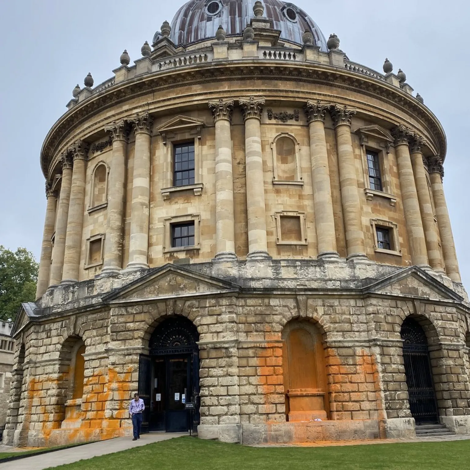 The Radcliffe Camera in Oxford shows orange paint splattered across its stone entrance arches and lower walls. The circular neoclassical building features ornate columns, a domed roof, and classical architecture. A person stands near the vandalized entrance, which appears to be cordoned off following the environmental protest.