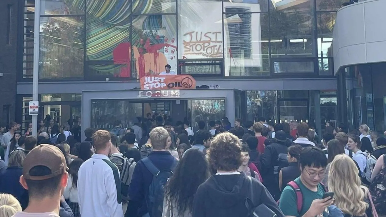 A large crowd of people, mostly young adults who appear to be students, gather outside a modern glass building. Orange 