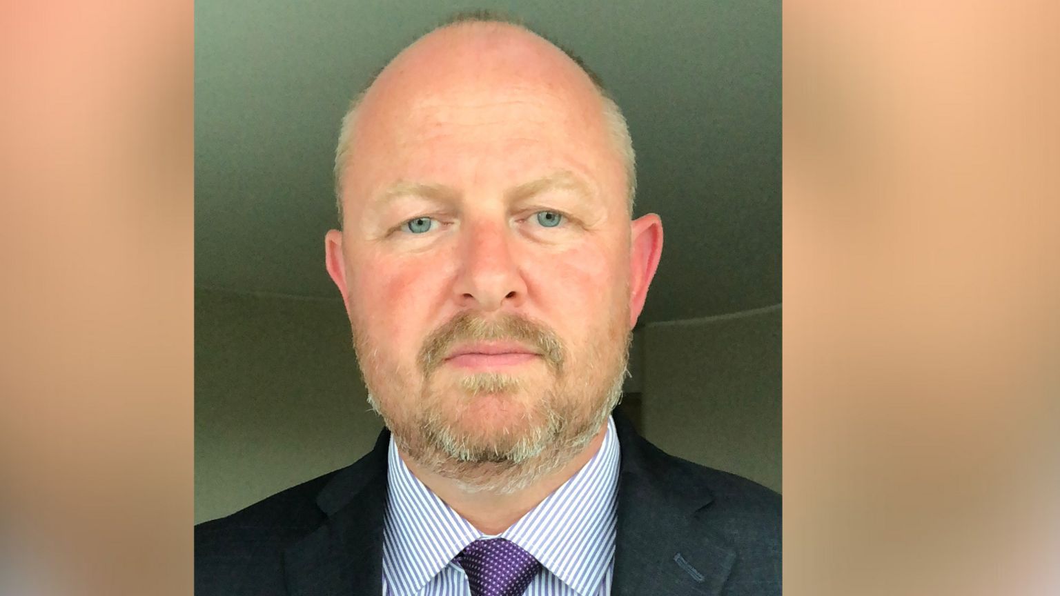 White man with purple tie and stripy shirt looks into camera