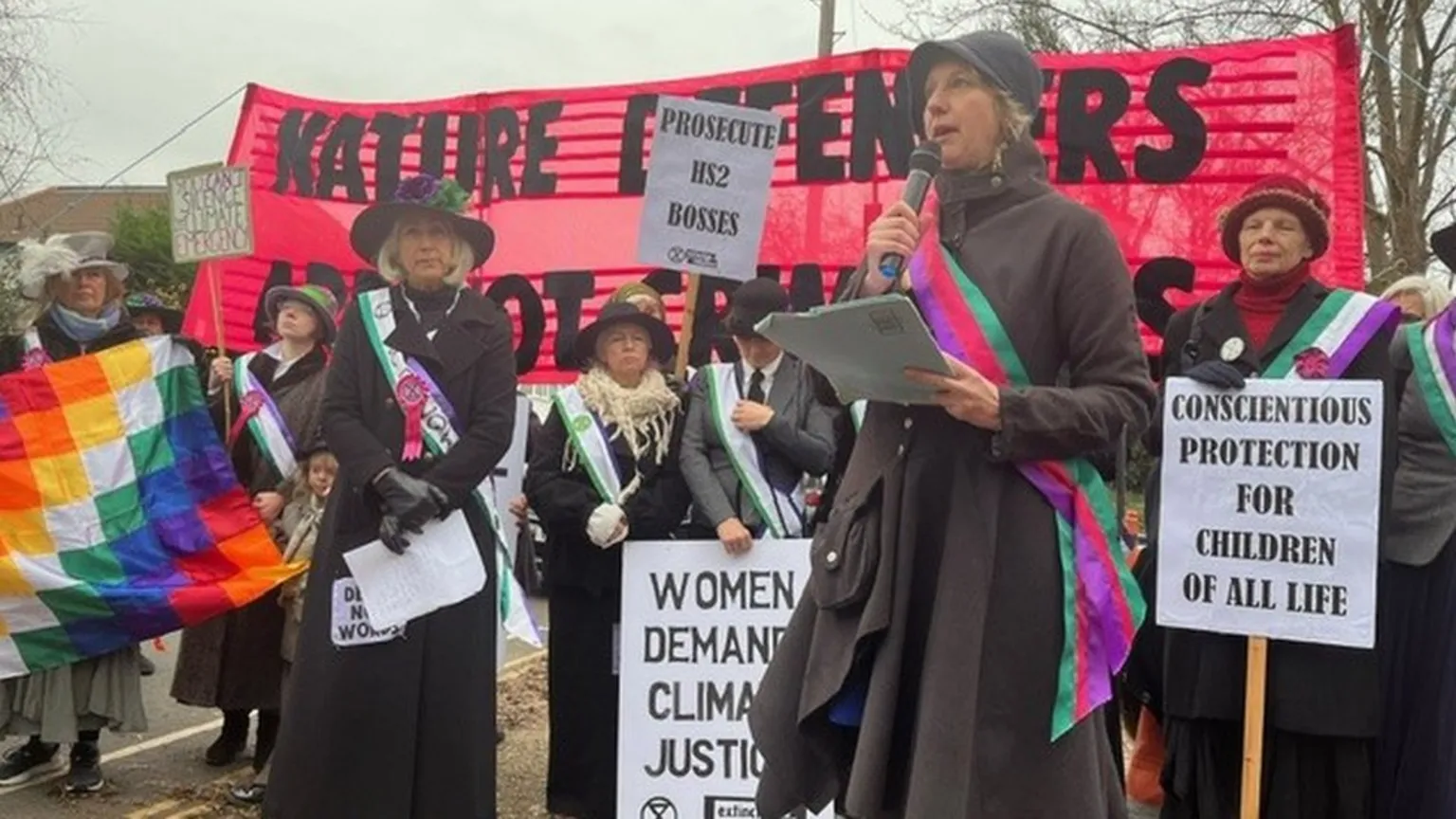 The image shows a woman in dark clothing speaking into a microphone at what appears to be a protest or demonstration. Behind her, supporters are dressed in period costume resembling suffragettes, wearing sashes and holding various signs including 