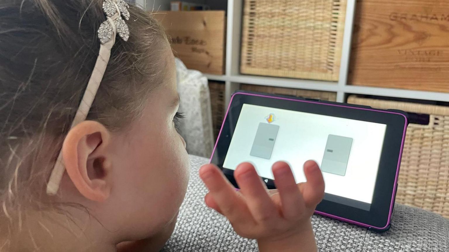 A young girl in a yellow dress and hairband is sitting on a couch and over her shoulder we can see a pink ipad with a game with two grey squares. Her hand is ready to touch the game. 