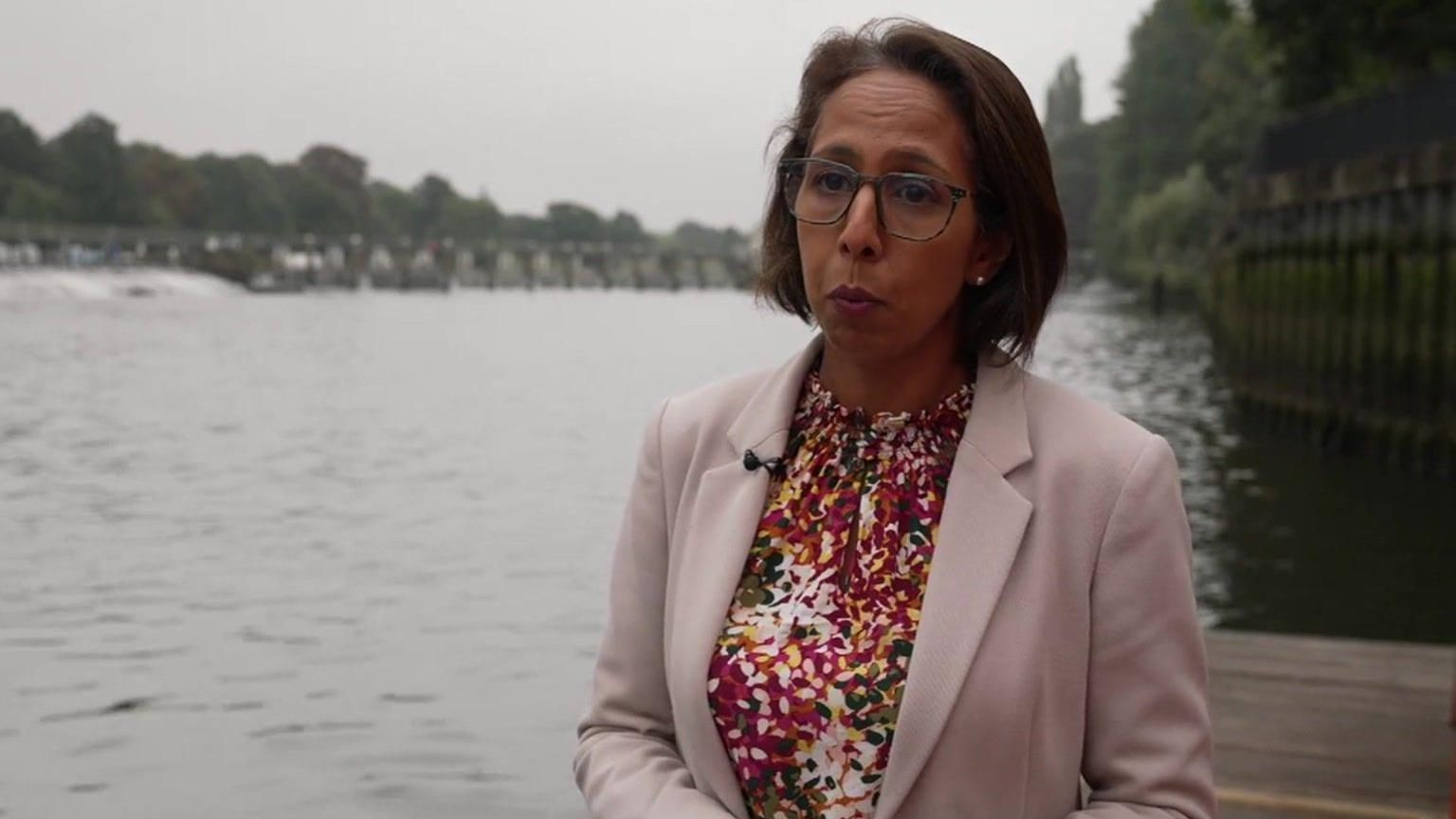 Munira Wilson standing along the River Thames in Teddington. She is wearing light pink blazer and floral top and has chin length bob and is wearing glasses.