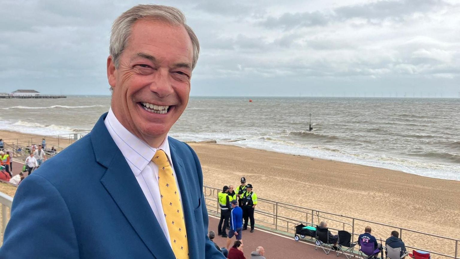 Nigel Farage smiling by the seafront in Clacton-on-Sea. He is wearing a navy suit and yellow tie