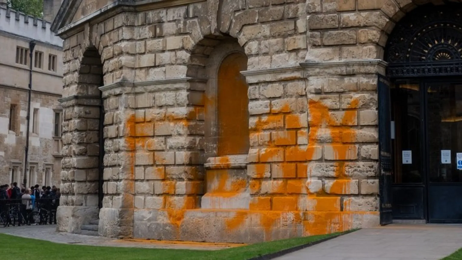 The image shows the historic Radcliffe Camera building in Oxford with bright orange paint splattered across its stone facade. The classical architecture features arched openings and rusticated stonework now covered in the distinctive orange paint associated with Just Stop Oil environmental protests.