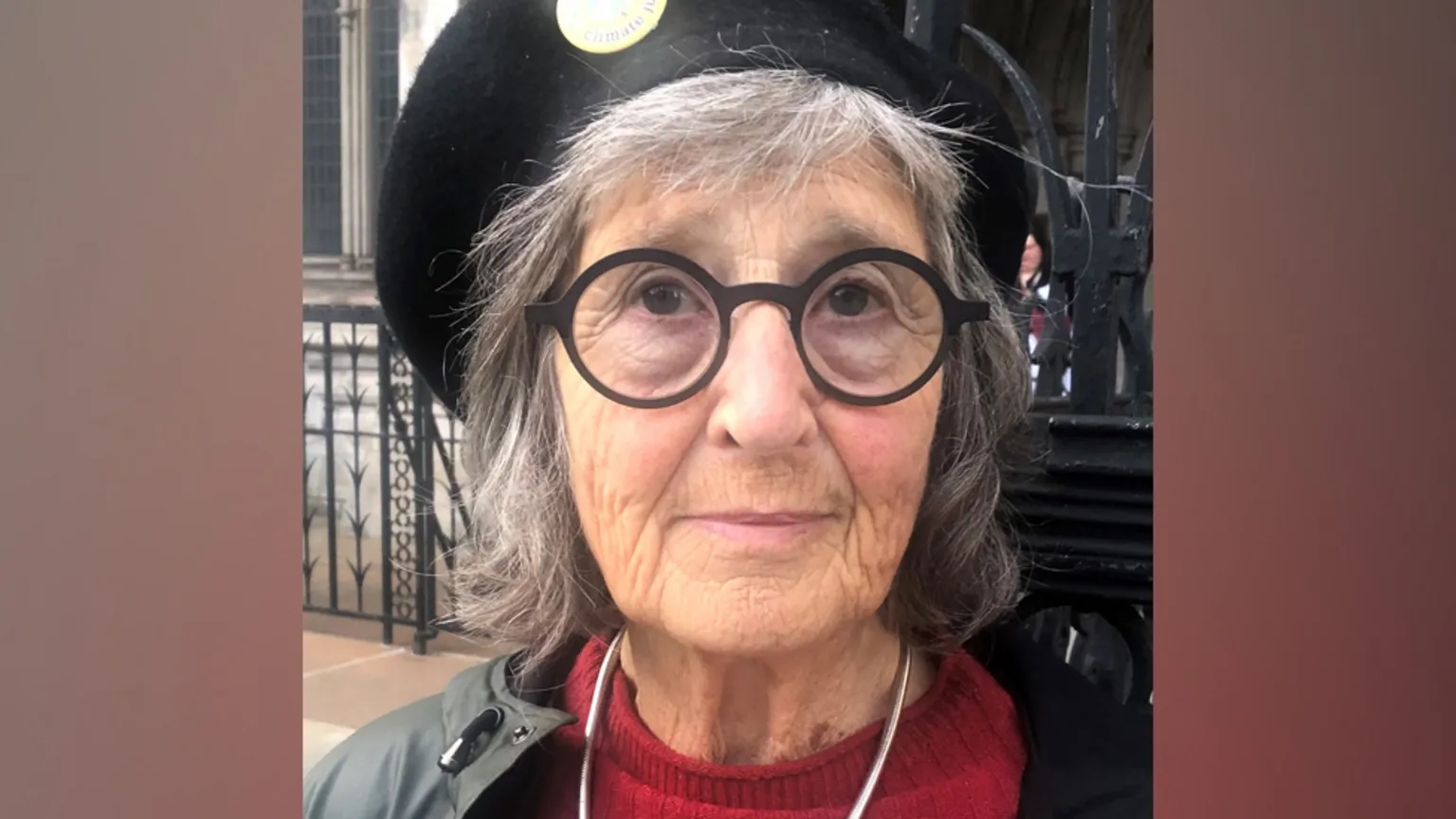 The image shows an elderly woman with gray hair wearing round black glasses and a red sweater. She has a slight smile and appears to be outdoors near a building with decorative ironwork visible in the background. She's wearing what appears to be a gray jacket or cardigan over her red top.