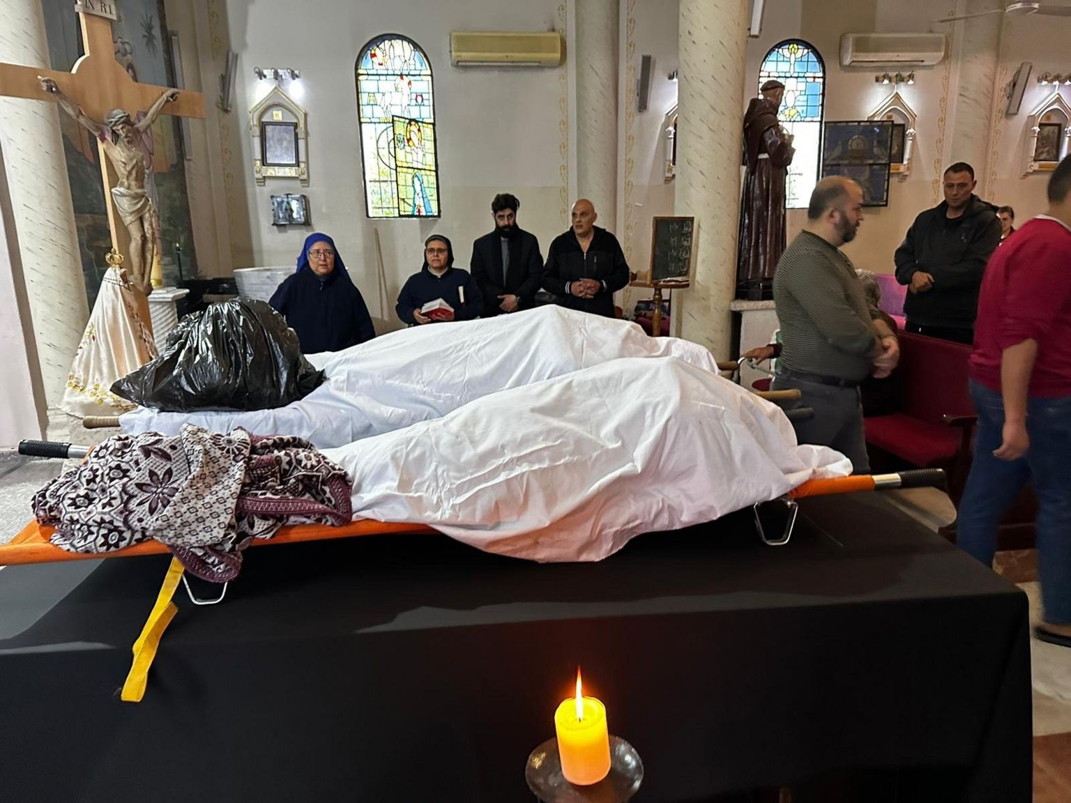 Two bodies covered in white sheets lay in a church, surrounded by people