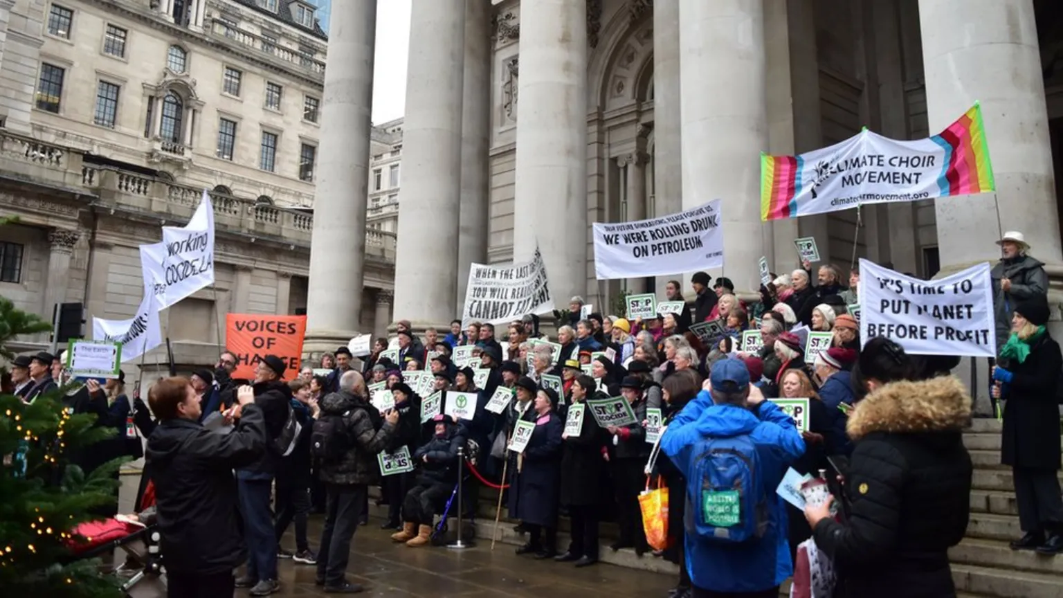 A large crowd of protesters gathers on the steps of a classical stone building with columns. They hold various banners including 