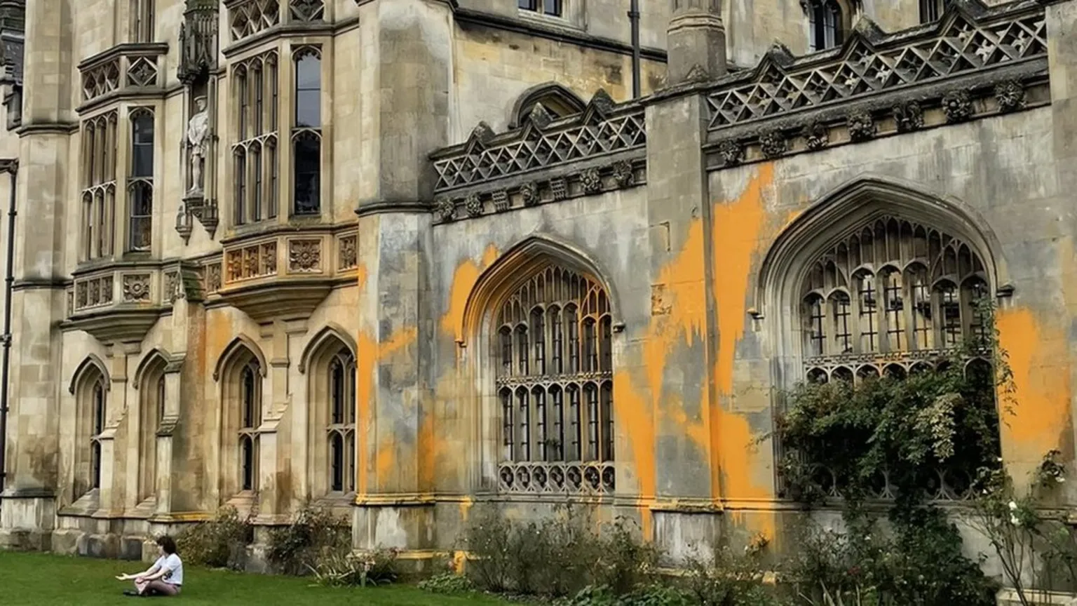 The image shows a Gothic stone building with ornate windows and architectural details covered in bright orange spray paint. A person sits on the grass in front of the vandalized building. The orange paint creates stark streaks across the historic stone facade.