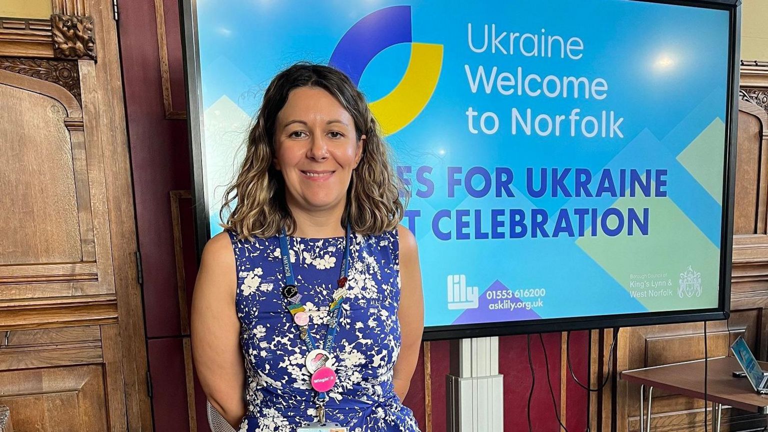 Rebekah Bensley-Mills has shoulder length hair. She is wearing a blue and white floral dress. She stands in front of a TV screen which says "Homes for Ukraine celebration" 