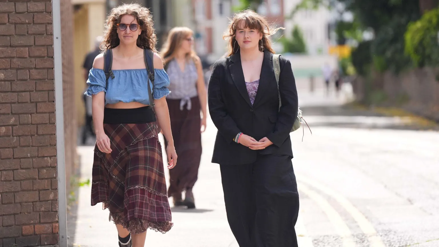 Two young women walk along a street, with one wearing a blue off-shoulder top, plaid skirt and glasses, while the other wears a black blazer over a patterned top. Both carry backpacks as they approach what appears to be a courthouse building with brick architecture visible in the background.