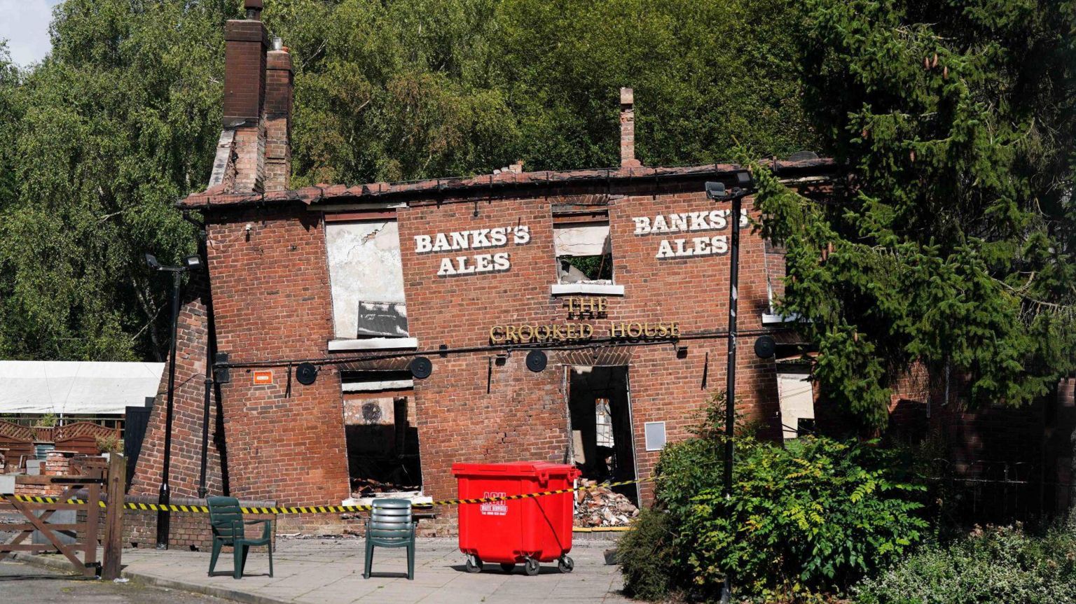 Crooked House pub shown before fire, leaning slightly to the left surrounded by black and yellow tape and with a skip outside.