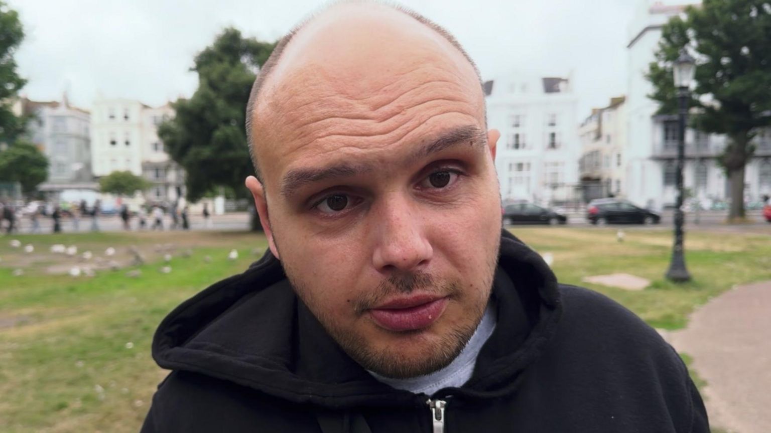 A close-up of Nick, who has a short beard and is wearing a grey t-shirt and black hoodie. He is standing in a park with a road and buildings visible behind it