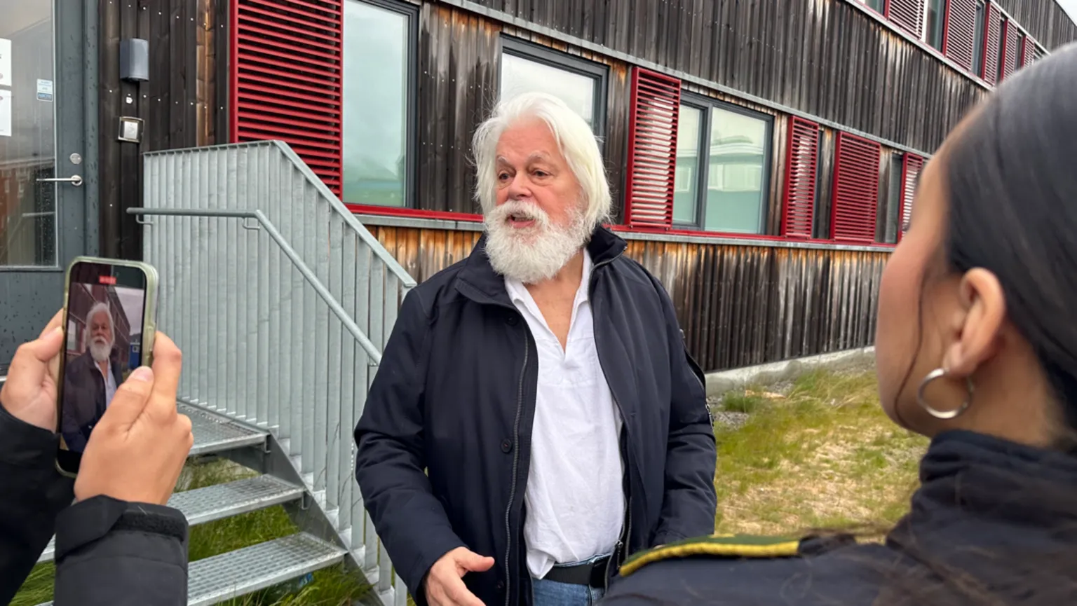 The image shows an older man with long white hair and a full white beard, wearing a dark jacket over a white shirt. He appears to be speaking outdoors near a modern building with red shutters. Someone is filming him with a smartphone, and another person is visible listening nearby.