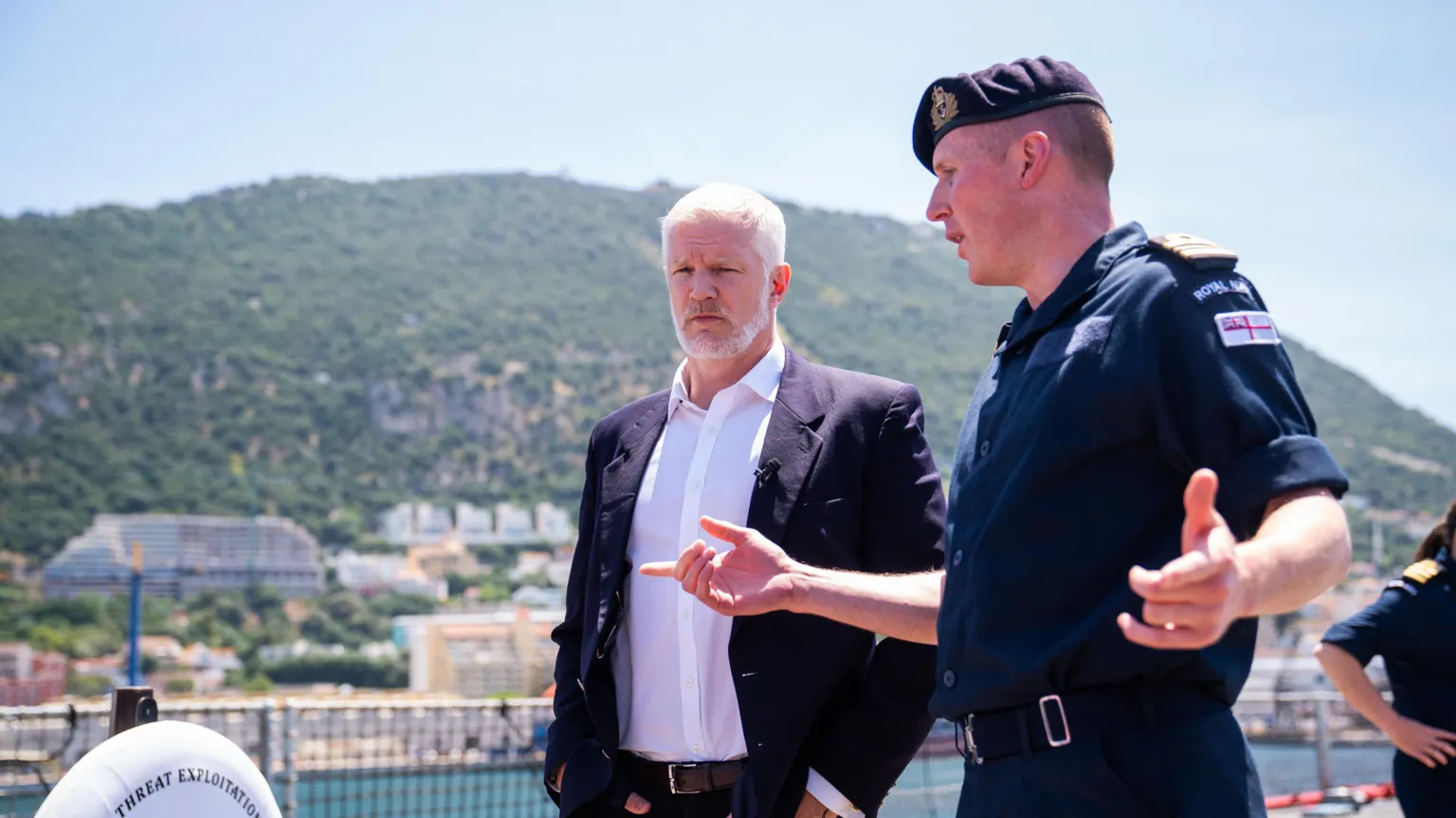 PA Media Two men, on the left is Al Carns wearing a dark suit jacket, trousers and brown belt, with a white shirt underneath. To his right is a man wearing a military uniform. They are pictured talking on an outside deck of a boat. In the distance, behind them there is a built up residential along the water and a mountain.
