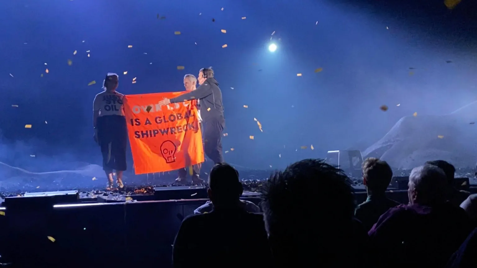 The image shows a stage performance being disrupted by protesters holding an orange banner that reads 
