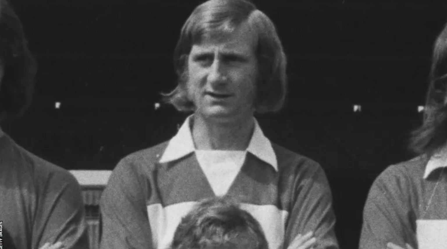 A black and white close up photo of Bill Gates posing for a team photo. He is wearing a striped polo football shirt and his hair falls below his ears. 