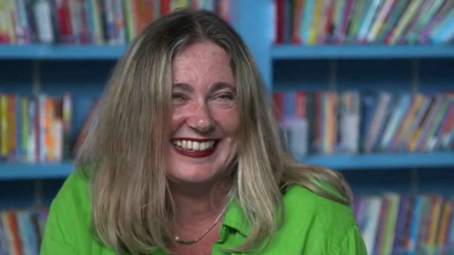 Headteacher Alison Bayliss with long blonde-brown hair wearing a green blouse, smiling with a bookcase in the background in soft focus