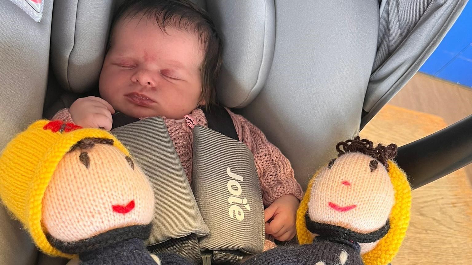 Baby Olive sleeping in a car seat, with two knitted firefighter toys next to her 