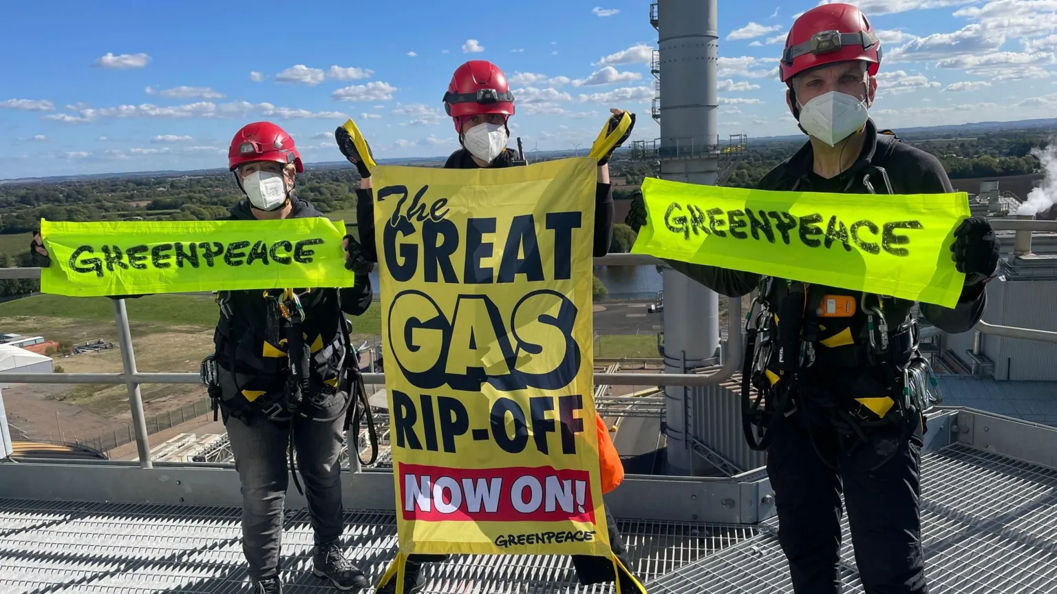 Three Greenpeace protesters wearing red helmets, face masks, and climbing gear stand on an industrial platform holding banners. Two hold bright green 