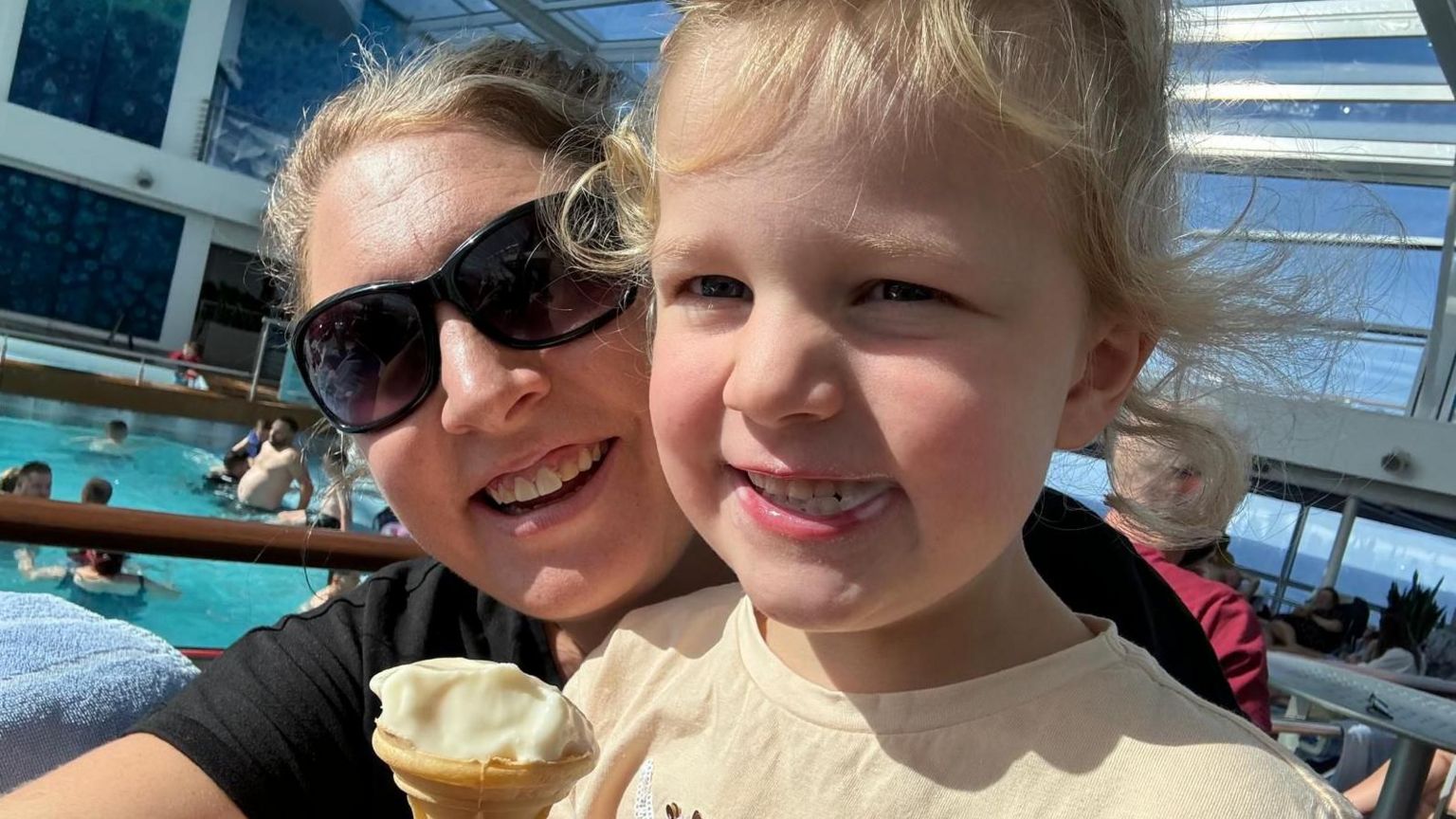 Megan Hall wearing sunglasses takes a selfie with her young daughter who is holding an ice cream with a pool behind them