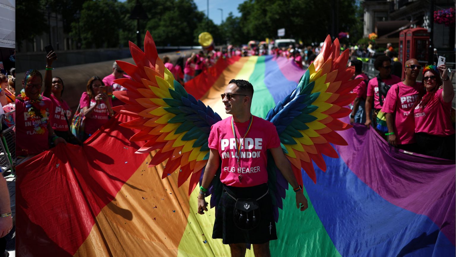 A parade flag bearer wearing a pink t-shirt and rainbow-coloured angel wings stands in front of a long rainbow flag. It stretches down the road behind him and is being held by dozens of people on each side