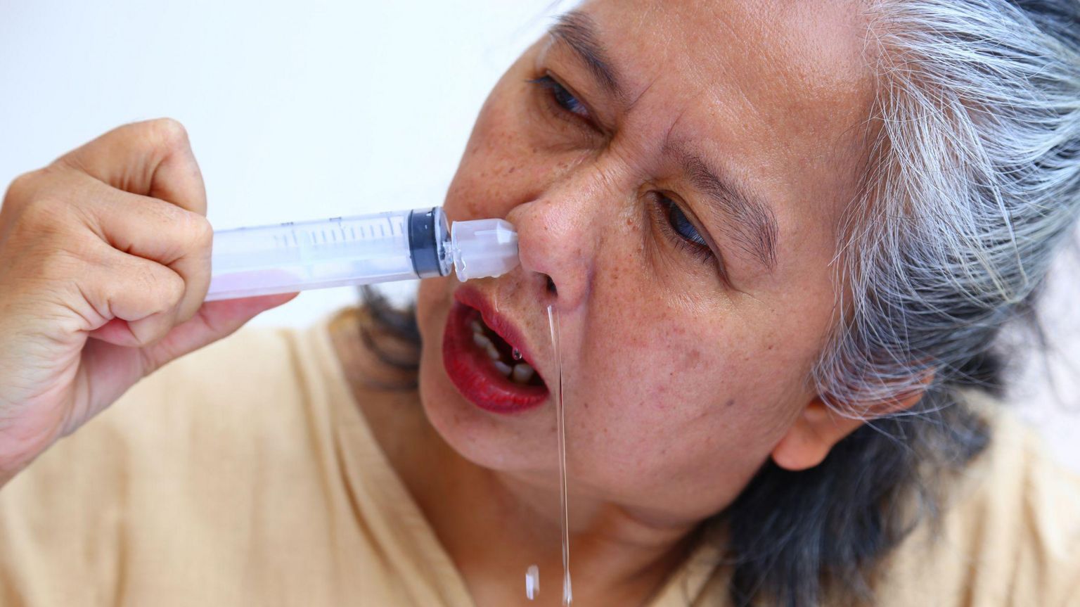 一名老妇将配备鼻腔冲洗喷嘴的注射器置于鼻孔内，水流从另一侧鼻孔流出。
