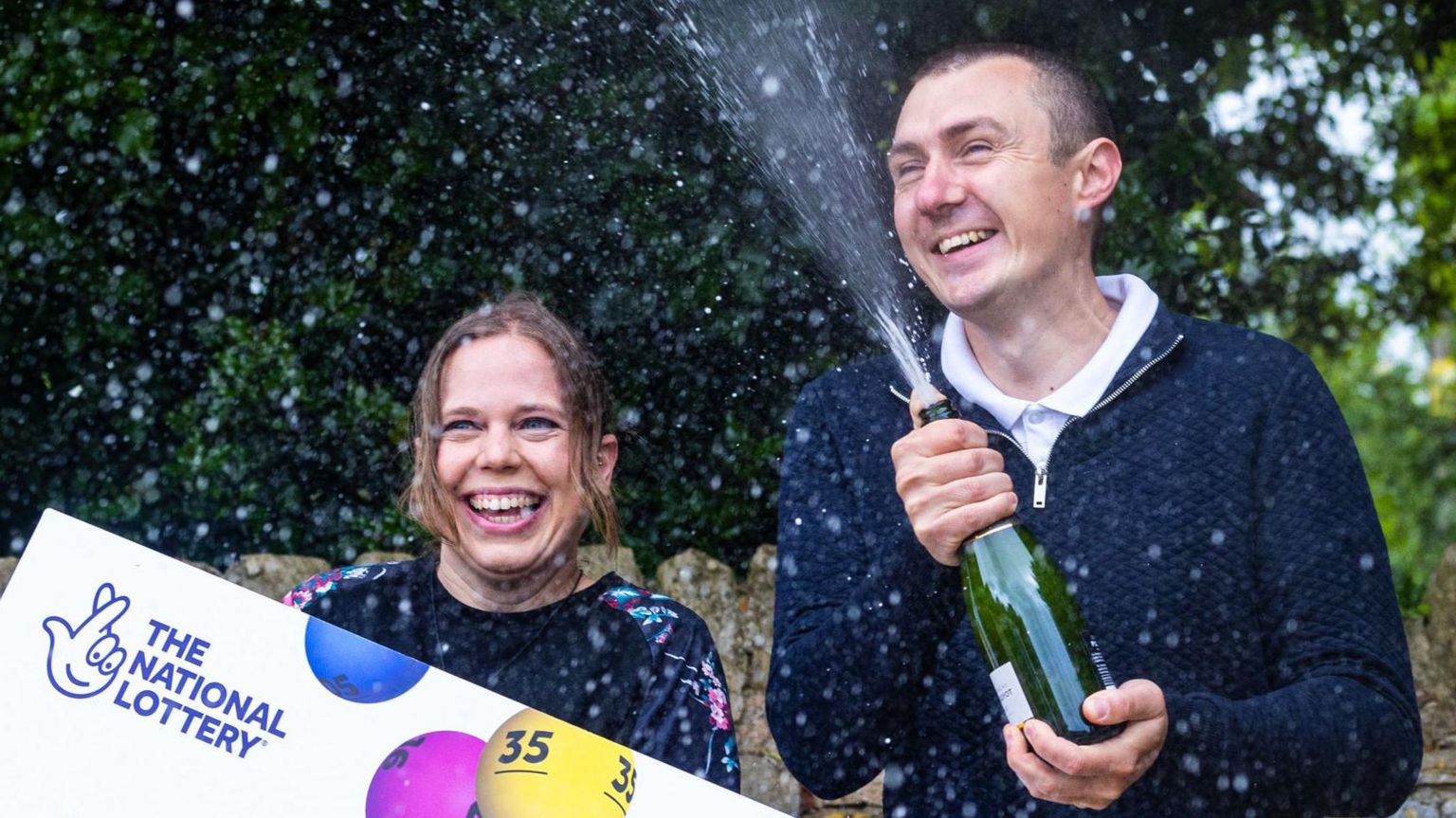 Katherine and Graeme White spraying fizzy wine from a bottle with a National Lottery sign