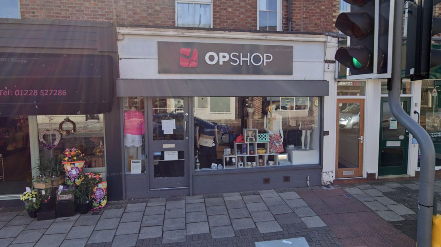 An exterior view of the OpShop charity shop. The shop on the high street has a grey, pink and white sign with clothes and bric-and-brac displayed in the window. There is a florist to the left.