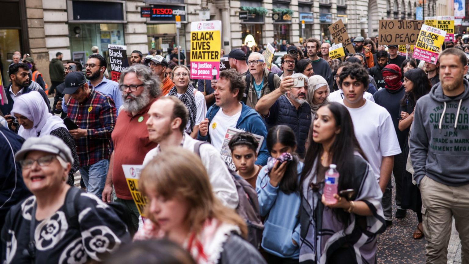 A group of people, some of whom are holding signs, including some that say 'No Justice No Peace' and 'Black Lives Matter' walk through the centre of Manchester