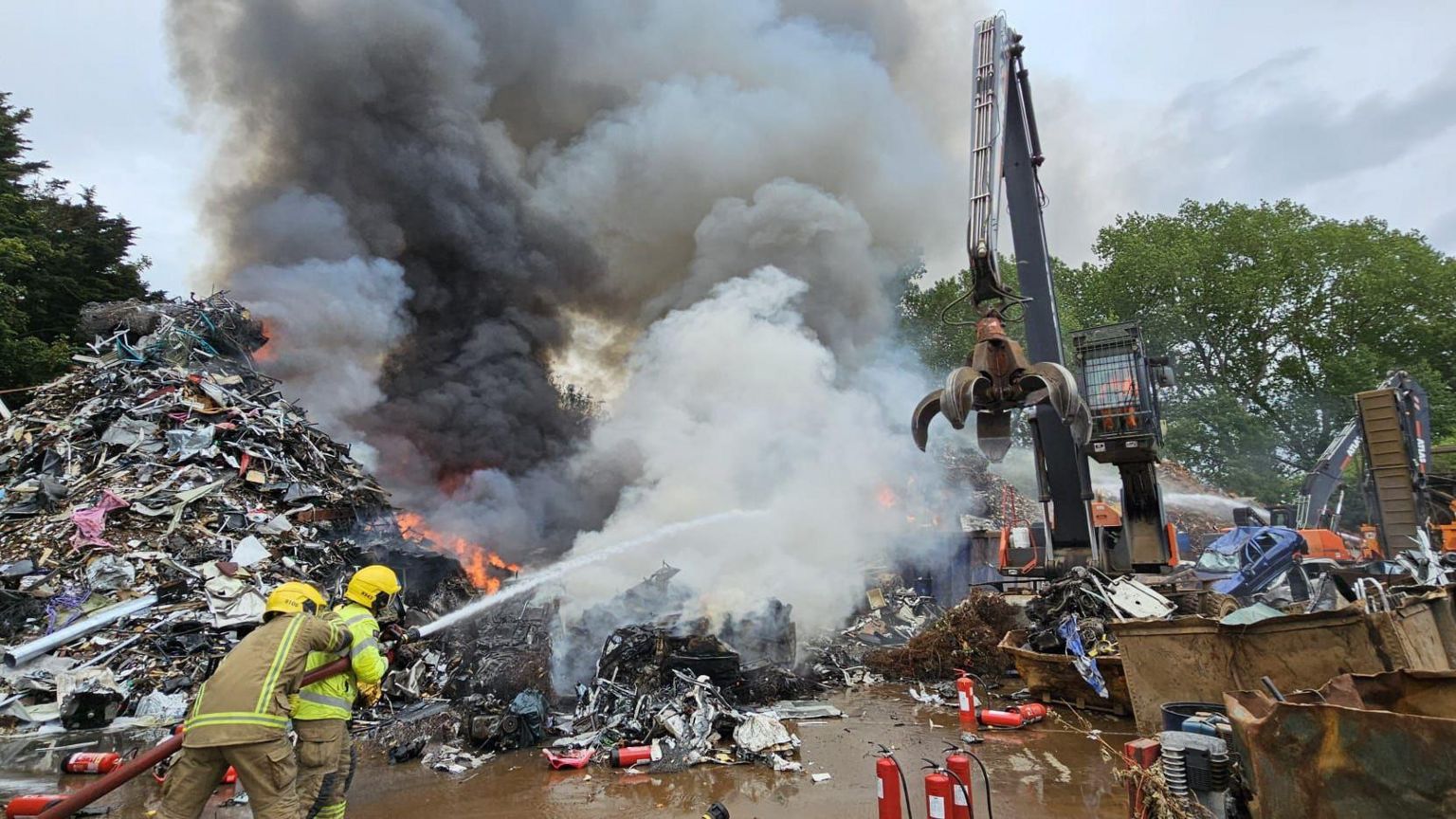 Two firefighters aim a hose at flames and smoke in a pile of scrap metal at John Huntley scrapyard