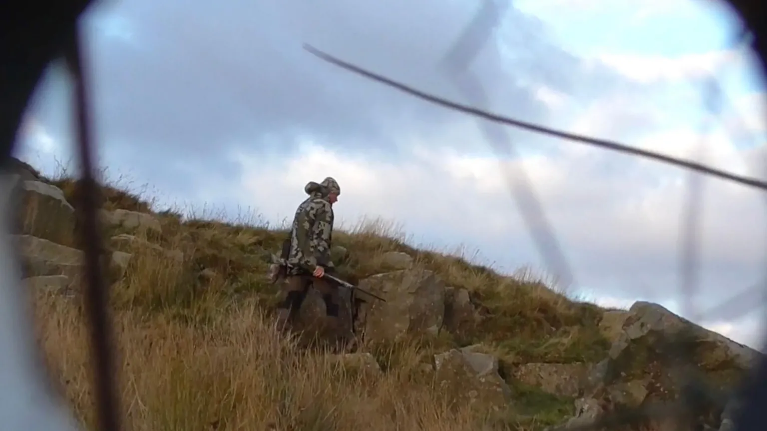 RSPB secret filming A man with a hood up on his head dressed in camouflage gear with a shotgun in his hands is walking across moorland. He is walking along some rocks and grassy land. Some black lines are visible in this screen grab from the covert footage and they are probably some grass in front of this covert camera set up for surveillance.