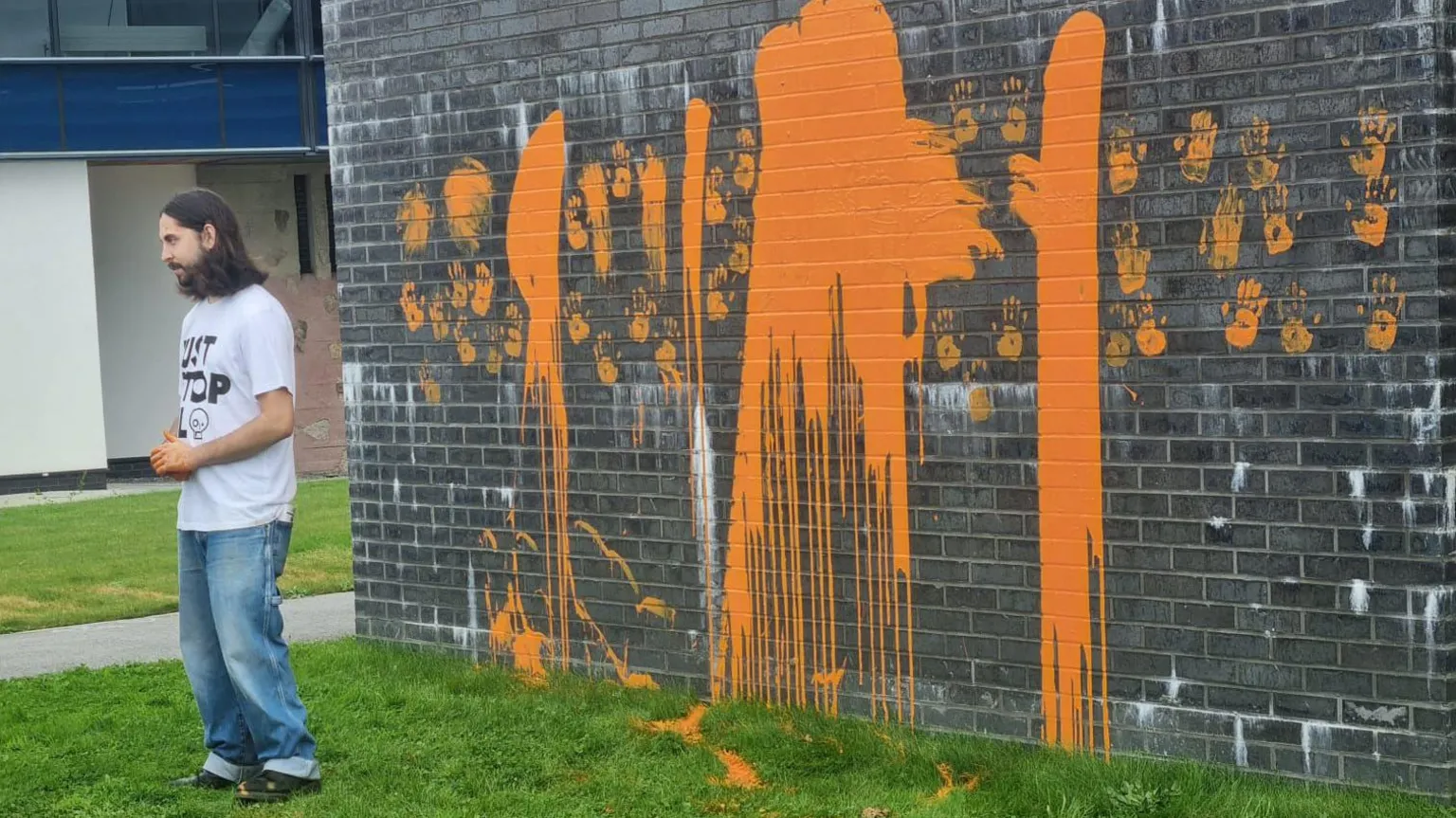 A man in a white t-shirt and jeans stands on grass beside a brick wall covered in orange paint. The paint appears to have been sprayed in large patches and drips across the dark brick surface. This occurred during a protest at Falmouth University's Penryn campus.