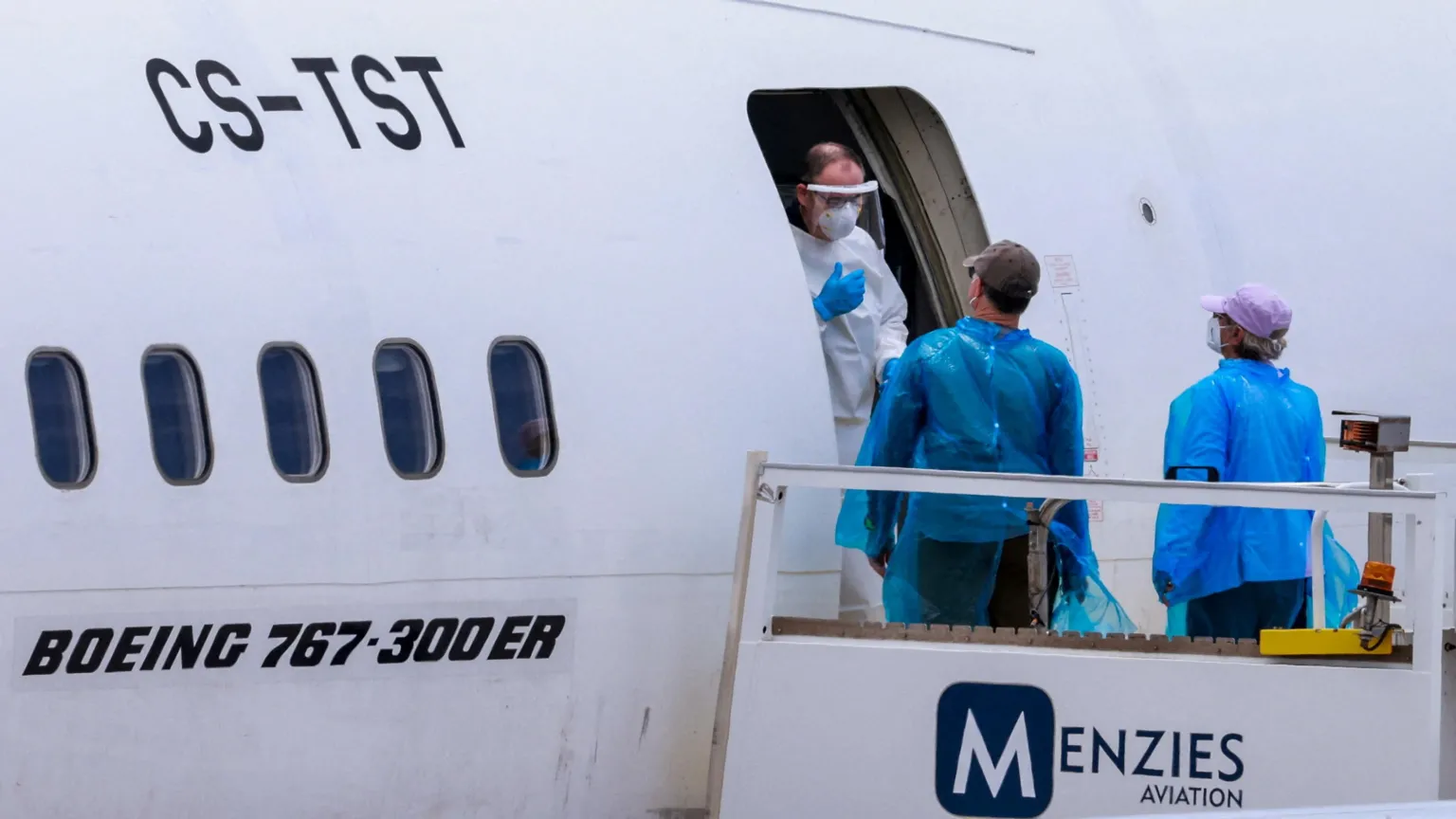 Reuters Man in full PPE talks to man and woman wearing blue plastic outer clothing and face masks at the entrance to a Menzies Aviation Boeing 767 plane.