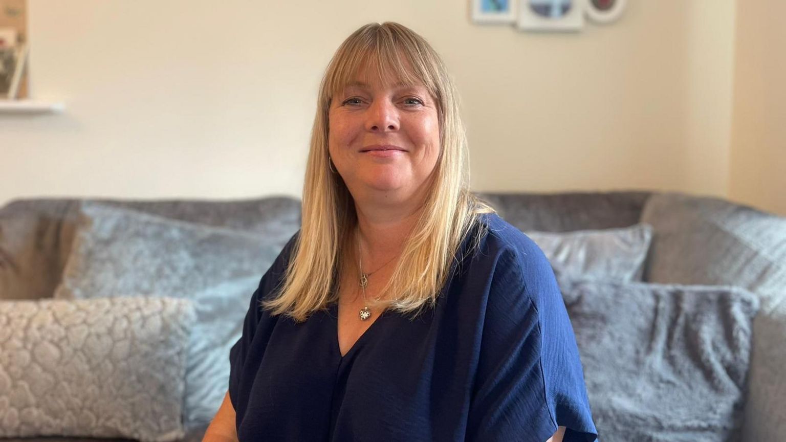 Sarah-Jane Clack sitting on a sofa wearing a blue top. She is blonde and smiling at the camera