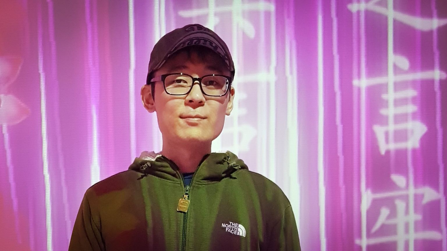 照片中，一名年轻的韩国男子张德俊戴着帽子，穿着深色连帽衫，背景是粉红色的。