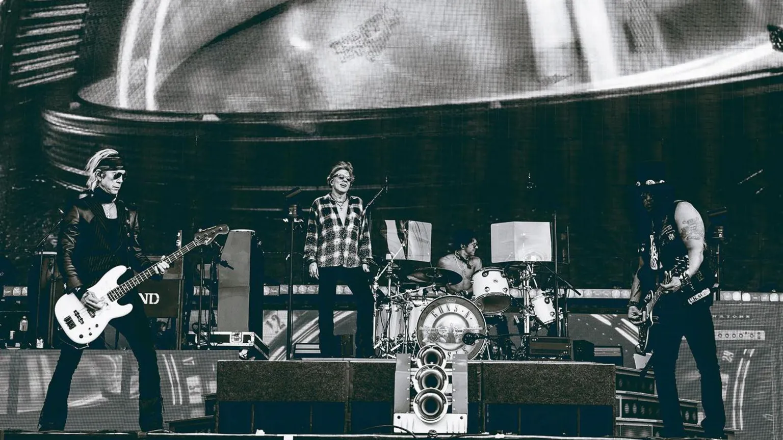 Guns N’ Roses A bass player, a man standing on a box and a guitarist on stage in a black and white photo.