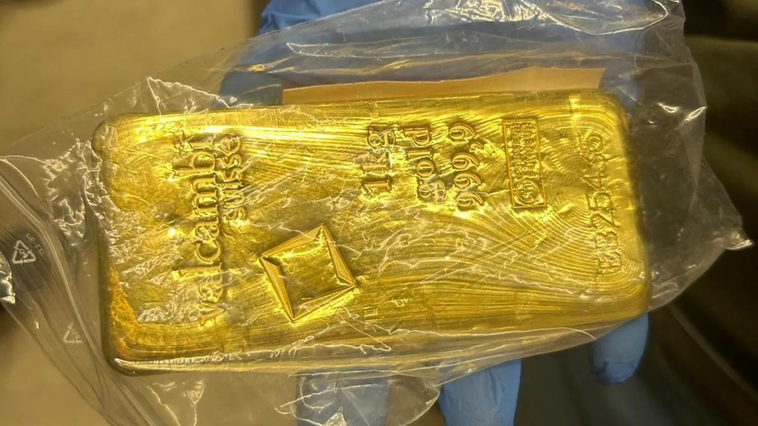 A person wearing blue gloves holding a bar of gold in a plastic bag. The bar has numbers, writing and shapes on it.