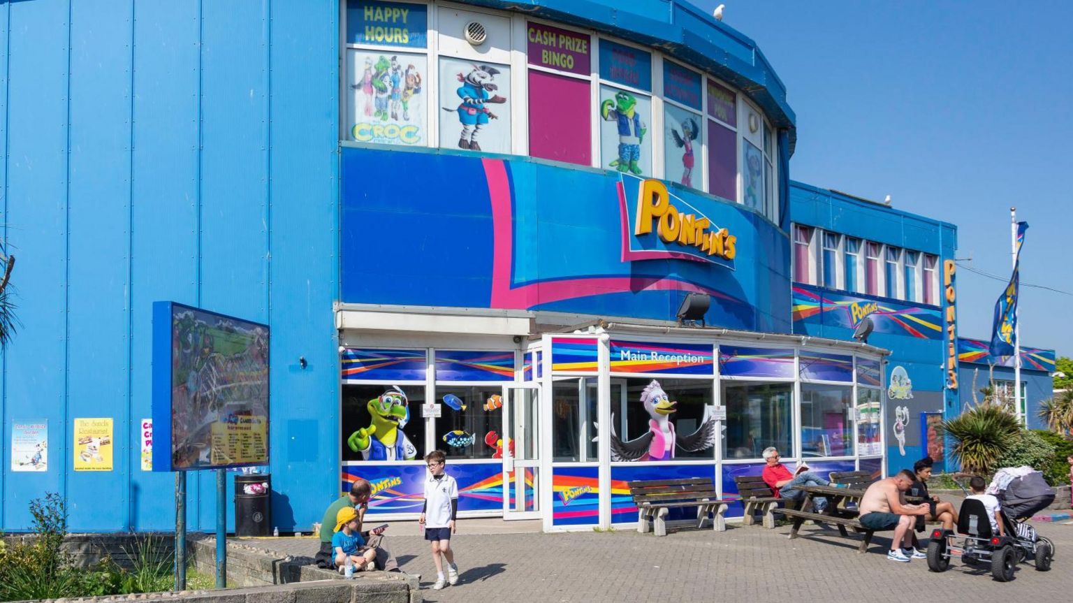 The exterior of a Pontins holiday park in the sun with several people standing or sitting in front of it