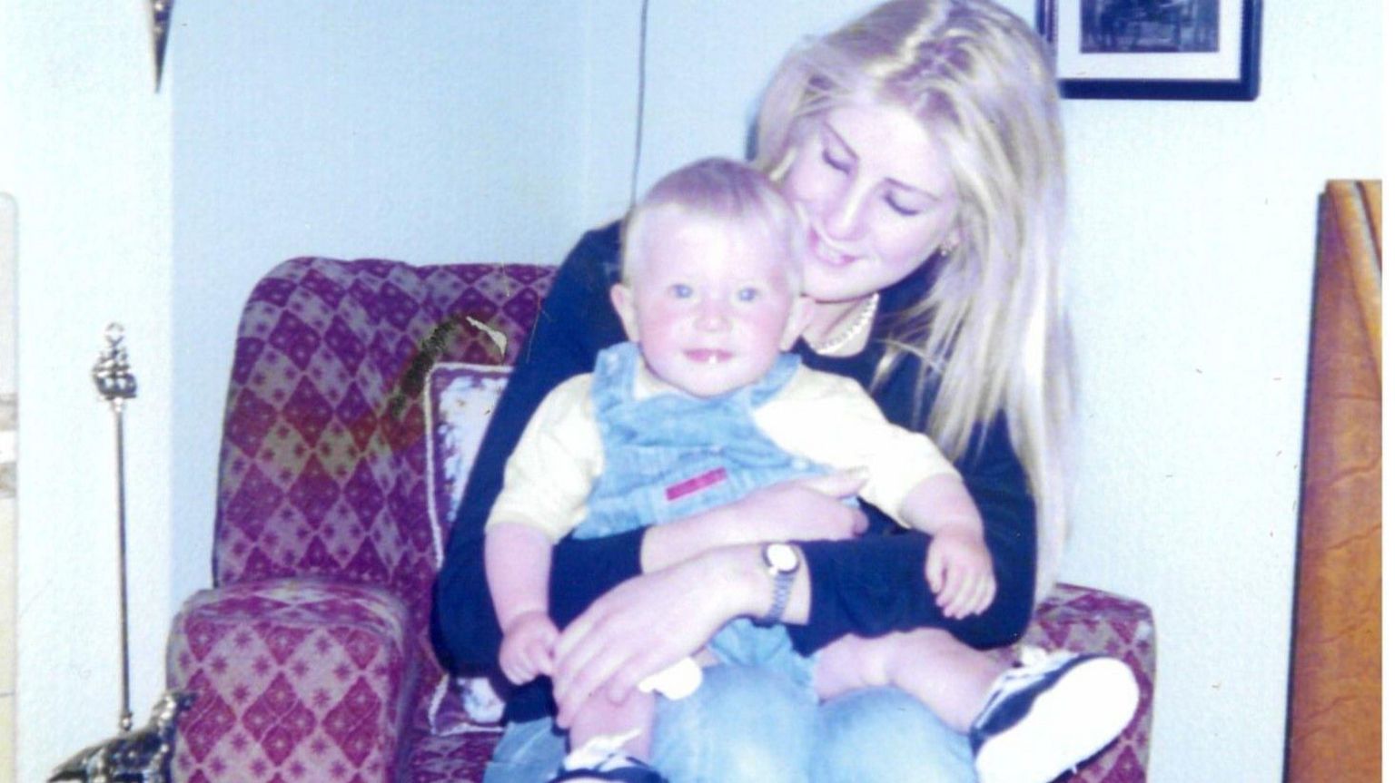 20-year-old Anna sits on an armchair holding her baby daughter Star. Anna has long blonde hair, while Star wears denim dungarees and smiles into the camera.