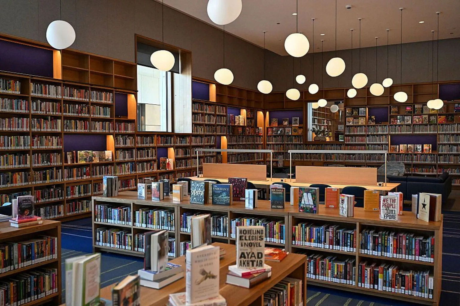 Joshua Lott for The Washington Post via Getty Images A room is lined with bookshelves and filled with book displayed made of light wood. There is a blue and green carpet and irregular orb lights hang