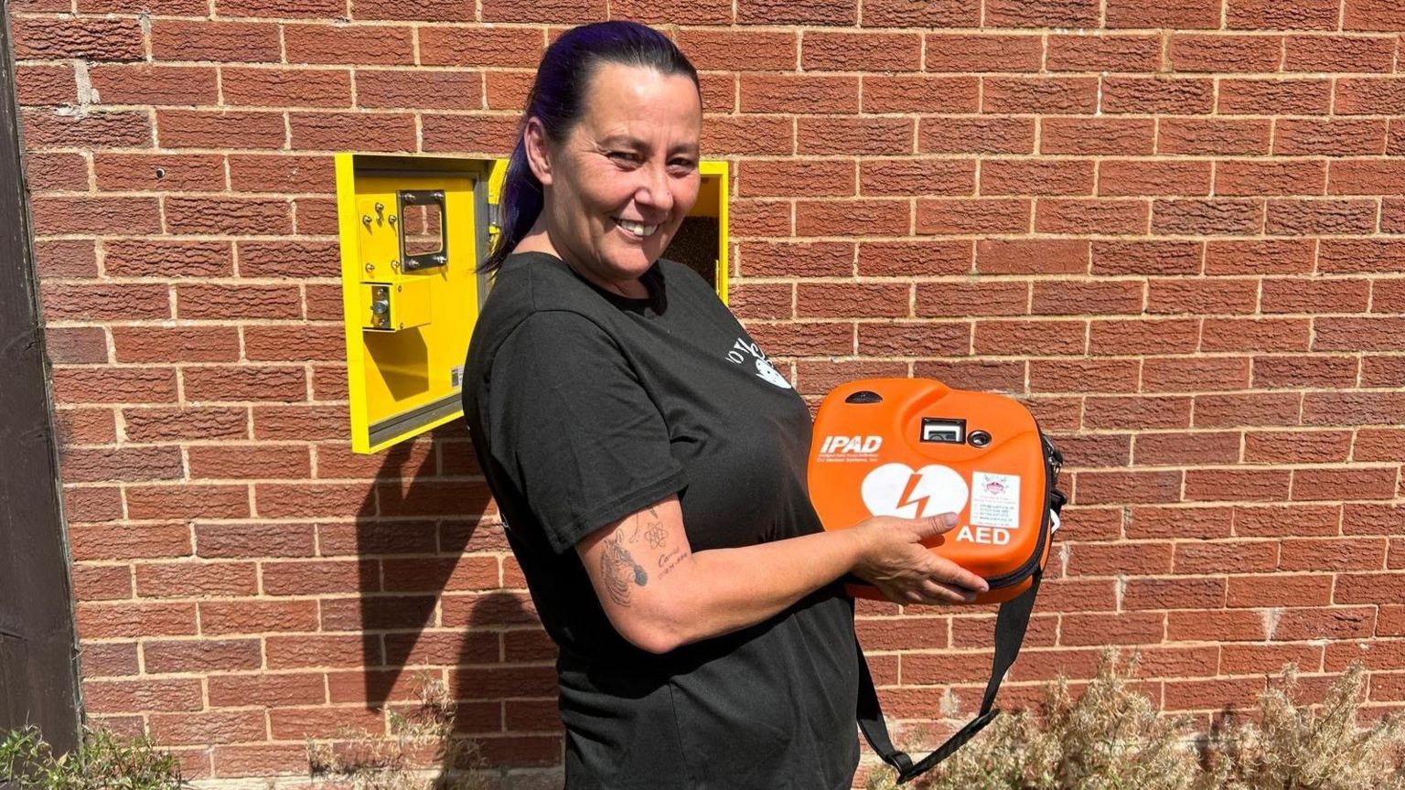 A woman is standing in front of a brick wall, holding an orange defibrillator