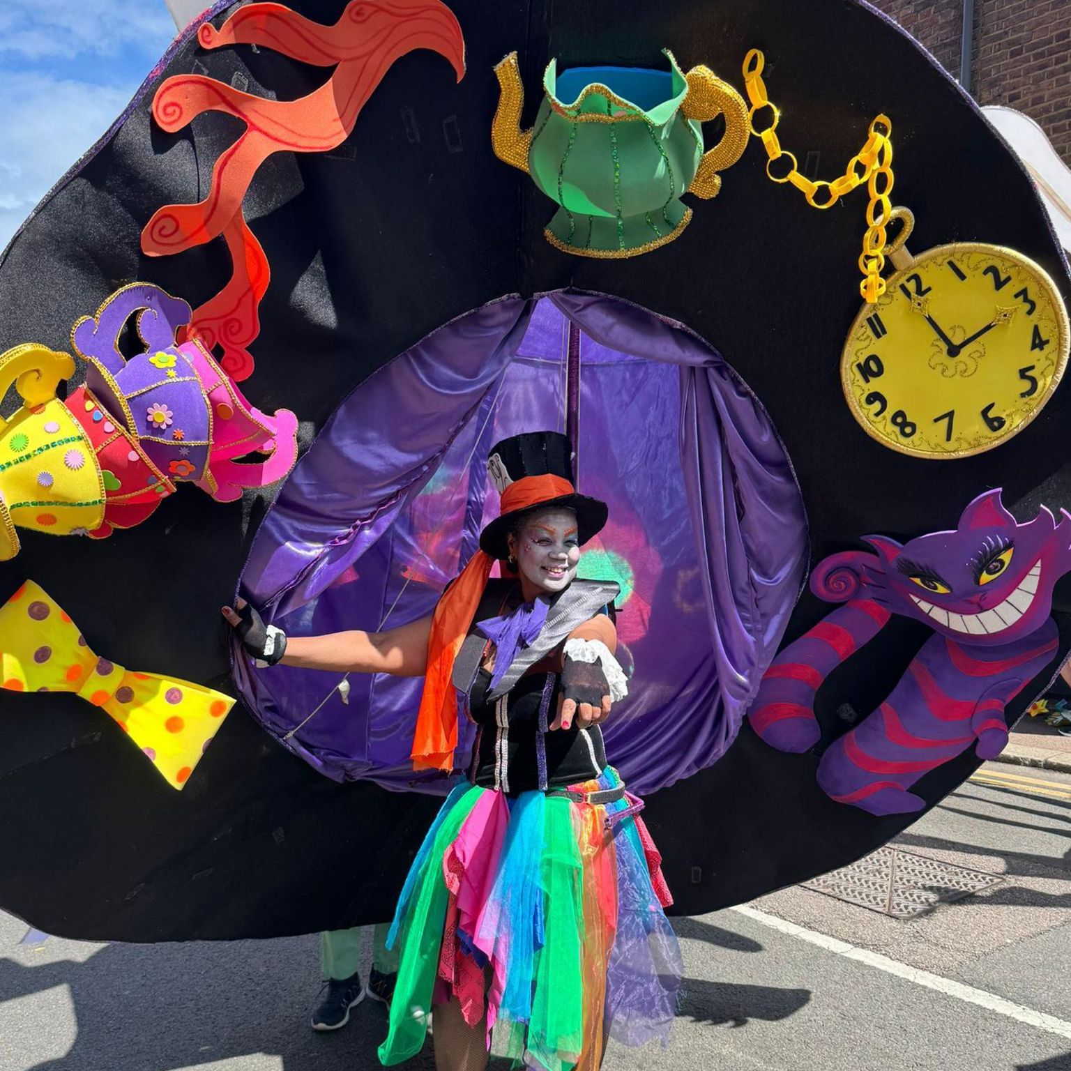 Maureen Drummond wearing an enormous hat costume which is three times her size, decorated with Alice in Wonderland characters and objects