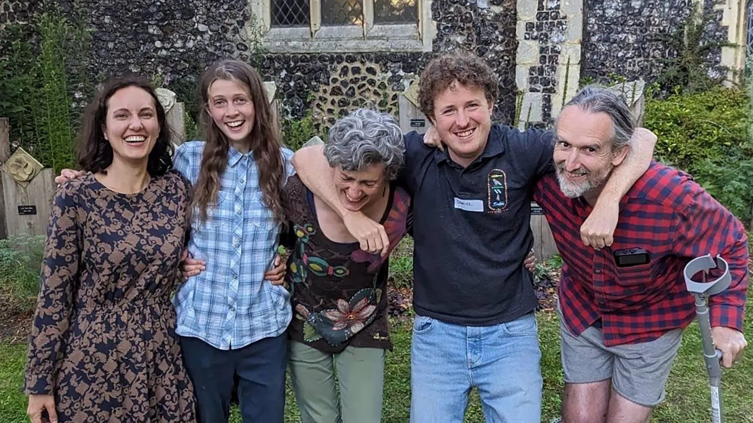 Five people stand together with arms around each other's shoulders, smiling at the camera in front of a stone building with flint walls. From left to right: a woman in a patterned dress, a woman in a blue checkered shirt, an older woman with gray hair, a young man in a dark polo shirt, and an older bearded man in a red plaid shirt holding a walking stick.