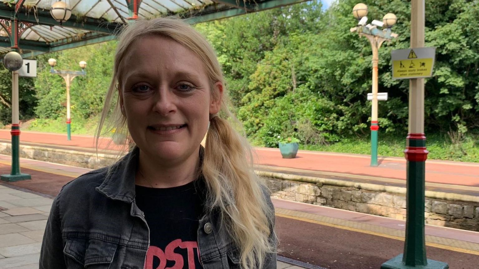 A blonde woman with long hair, wearing a denim jacket and black t-shirt, standing on a railway platform.