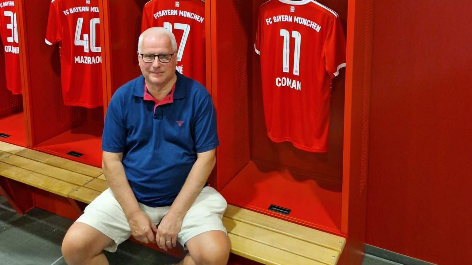 Mike Ash sitting on a bench in a changing room wearing a blue shirt and white shorts. Behind him are cubby holes with red football shirts hanging on  hooks