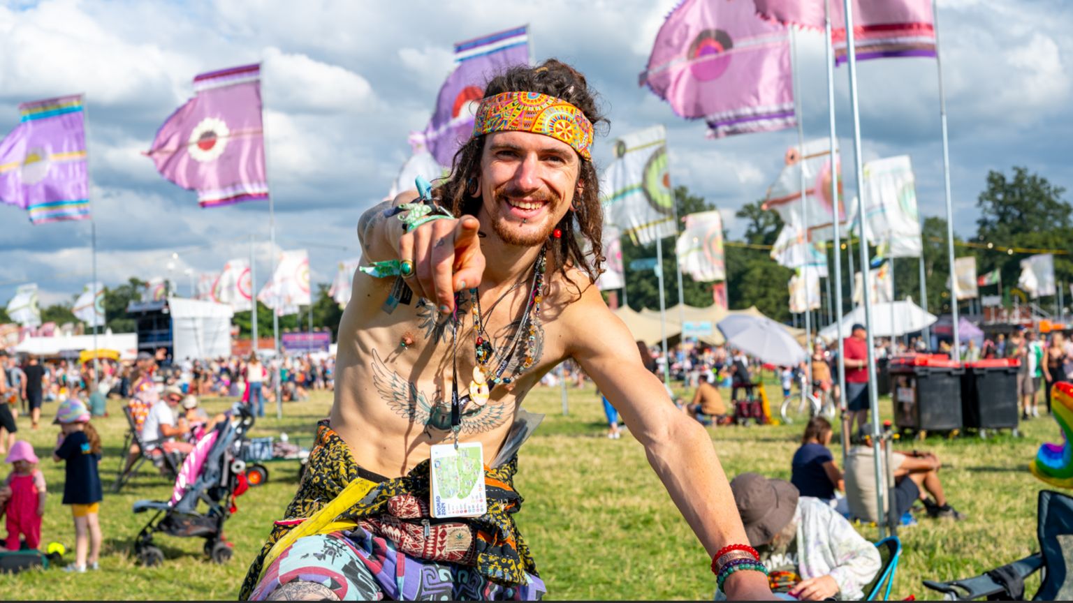 A person wearing a colourful bandana, dreadlocks with beads around their neck and a tattoo across their chest pointing at the camera