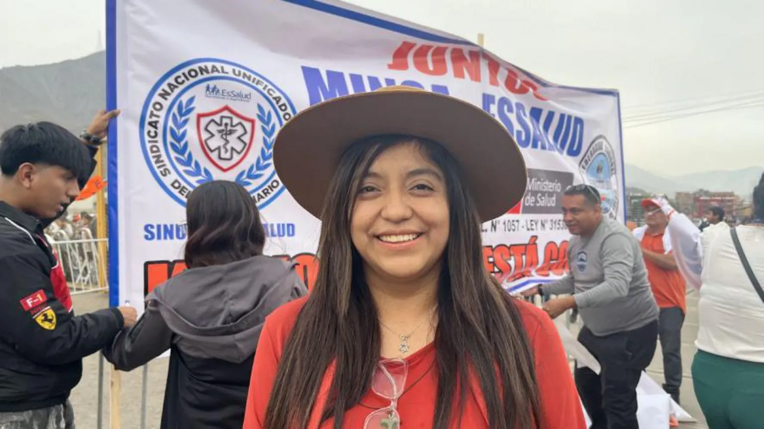 Janeth wearing a red dress and top, with long hair seen under a hat. She is standing in front of a campaign poster.