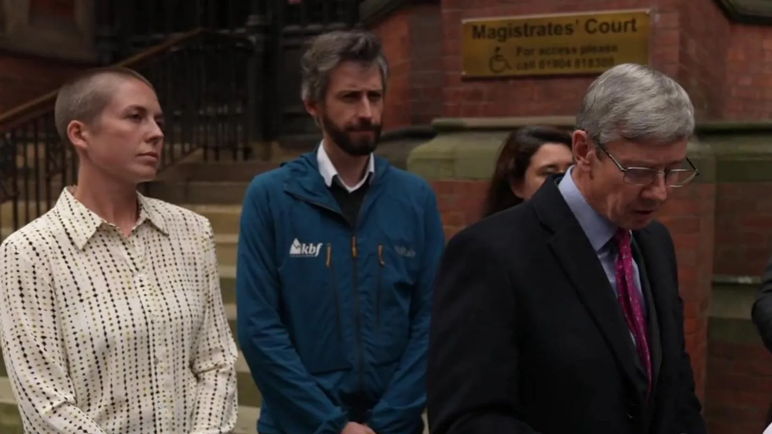 The image shows people standing outside a Magistrates' Court building. On the left is a person with short hair wearing a white polka-dotted shirt. In the center is a bearded man in a blue jacket. On the right is an older man in a dark suit with glasses and a red tie, looking downward.