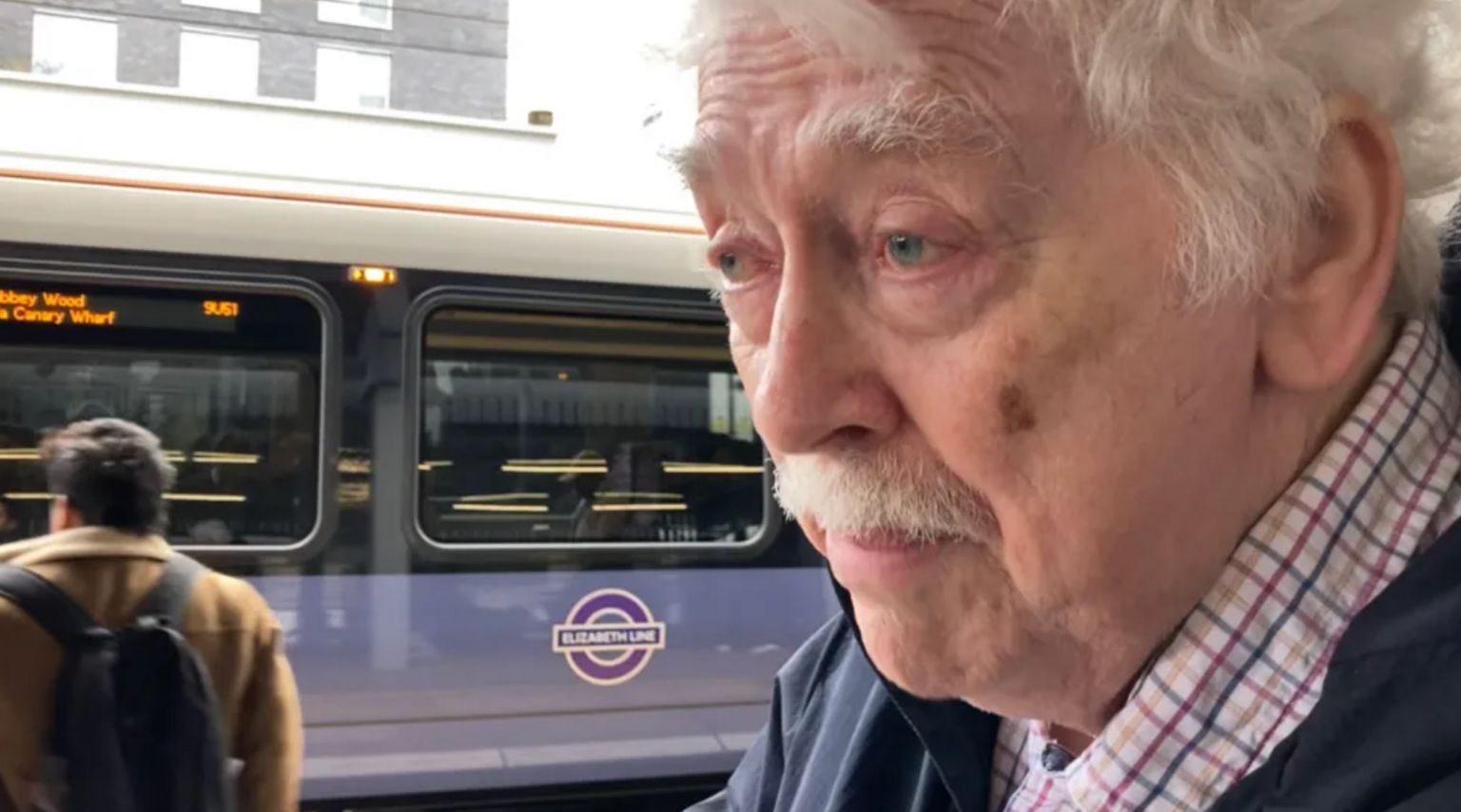 Eric Leach is standing on a train platform in front of an Elizabeth Line Tube train. He has a moustache and a checked shirt