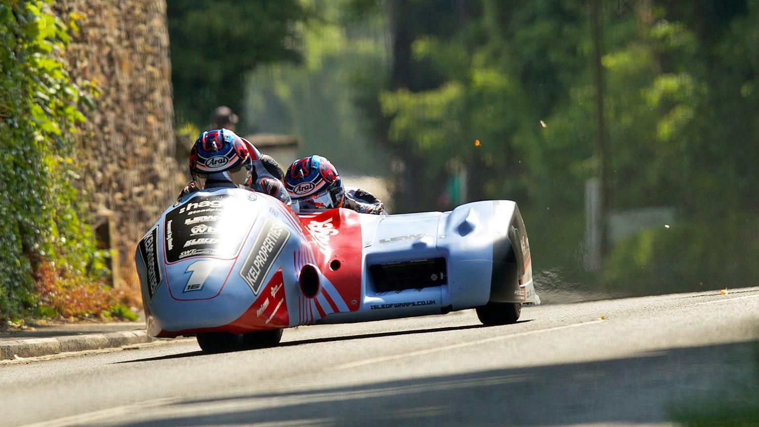 A light blue and red sidecar outfit on a race track with hedges and greenery in the background